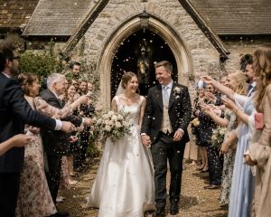A bride and groom walking hand in hand through confetti thrown by guests outside a stone English church.